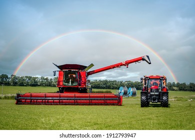 Brand New Modern Clean Combine Harvester And Shiny Red Agricultural Tractor In Rural Farm Field On Summers Day With Beautiful Colourful Rainbow In The Sky Behind