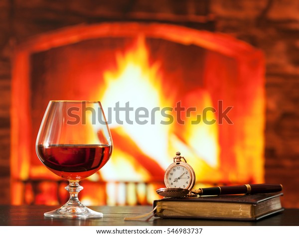 Brandy glass, diary and pocket watch on the table near the burning fireplace
