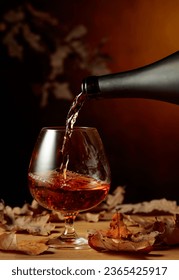 Brandy is poured from a bottle into a glass. A snifter of brandy on an old oak table with dried-up oak leaves. - Shutterstock ID 2365425917