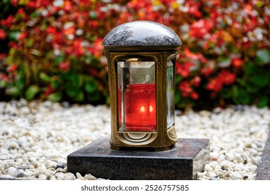 a brass grave lantern in a cemetery with a burning red candle on a marble pedestal amidst white pebbles in front of red plants in a blurred background - Powered by Shutterstock