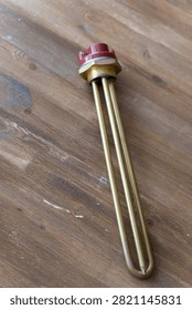 Brass heating element rests on wooden surface. Red cap seals electrical connection point. Simple industrial design for water or liquid heating. Ideal for plumbing, heating, or boiler system visuals