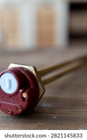 Brass heating element rests on wooden surface. Red cap seals electrical connection point. Simple industrial design for water or liquid heating. Ideal for plumbing, heating, or boiler system visuals