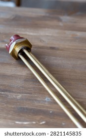 Brass heating element rests on wooden surface. Red cap seals electrical connection point. Simple industrial design for water or liquid heating. Ideal for plumbing, heating, or boiler system visuals