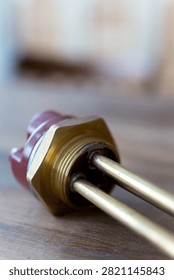Brass heating element rests on wooden surface. Red cap seals electrical connection point. Simple industrial design for water or liquid heating. Ideal for plumbing, heating, or boiler system visuals