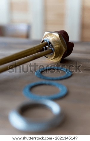 Brass heating element rests on wooden surface. Red cap tops the metallic tube. Three gaskets sit nearby - two blue, one silver. Tools lie still, unattended, on rustic wood. Ideal for plumbing, heating