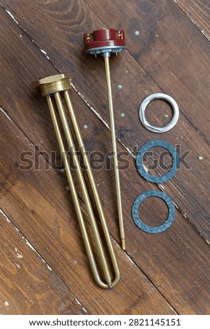 Brass heating element rests on wooden surface. Red cap tops the metallic tube. Three gaskets sit nearby - two blue, one silver. Tools lie still, unattended, on rustic wood. Ideal for plumbing, heating