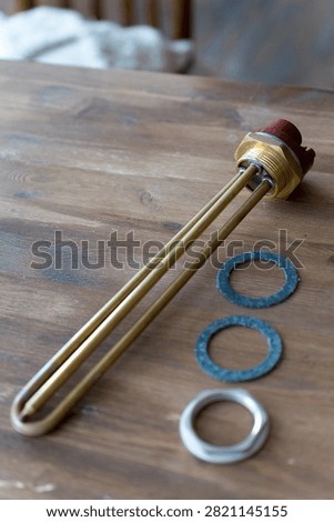 Brass heating element rests on wooden surface. Red cap tops the metallic tube. Three gaskets sit nearby - two blue, one silver. Tools lie still, unattended, on rustic wood. Ideal for plumbing, heating