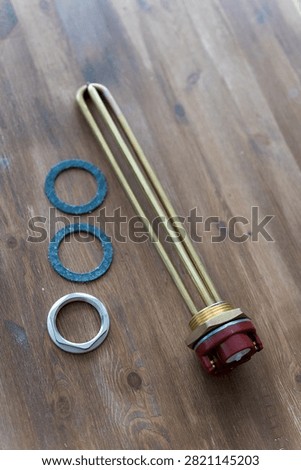 Brass heating element rests on wooden surface. Red cap tops the metallic tube. Three gaskets sit nearby - two blue, one silver. Tools lie still, unattended, on rustic wood. Ideal for plumbing, heating