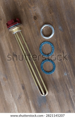 Brass heating element rests on wooden surface. Red cap tops the metallic tube. Three gaskets sit nearby - two blue, one silver. Tools lie still, unattended, on rustic wood. Ideal for plumbing, heating