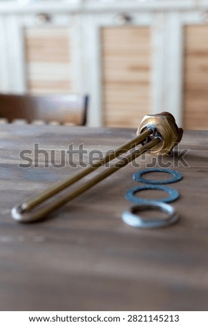 Brass heating element rests on wooden surface. Red cap tops the metallic tube. Three gaskets sit nearby - two blue, one silver. Tools lie still, unattended, on rustic wood. Ideal for plumbing, heating