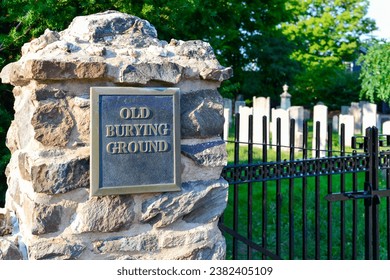 A brass metal sign with old burying ground on a rock gate post entrance to a graveyard. The black metal fence surrounds the cemetery. The headstones in the burial site are made of white stones.  