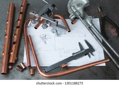 Brass pipes, pipe bender, pipe rammer tool and a layout drafts on a fitter workbench.