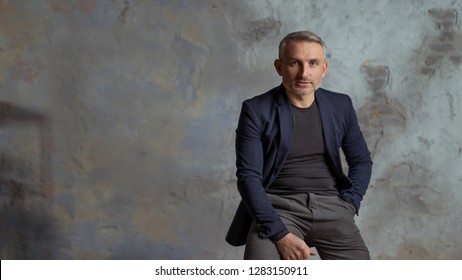 Brave 45 Years Old Men With Gray Hair And Beard Sits On The Chair, Gray Background