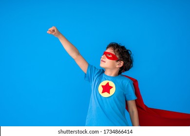 Brave Boy In A Superhero Costume, Wearing A Red Cape And Mask, Held Out His Hand As A Sign Of Desire To Win, Isolated On A Pink Background. Cute Kid Playing Superhero. The Concept Of Power And Justice