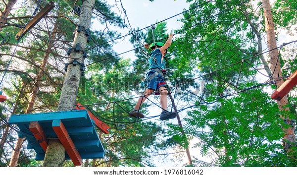 A brave boy walks along a rope ladder stretched\
between trees at a great height. Family fun in the adventure high\
rope park. Extreme kinds of entertainment for children during\
summer holidays.