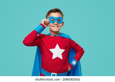 Brave cheerful boy in superhero cape and mask standing with hand on waist on blue background and looking away - Powered by Shutterstock