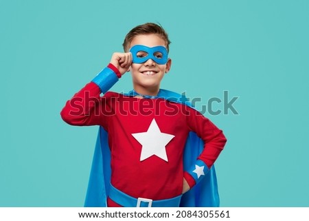 Brave cheerful boy in superhero cape and mask standing with hand on waist on blue background and looking away