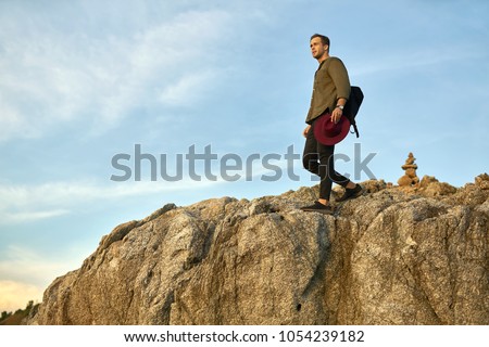 Brave guy with a black backpack walks on the rocky cliff on the sky background. He wears dark jeans and sneakers, olive shirt and holds a crimson hat. Man looks to the side. Sun shines onto his body.