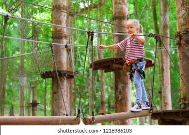 Brave Little Child In Safety Harness Climb On Tree Tops Route To Zip Line, Pass Hanging Bridge Obstacle In Adventure Rope Park. Outdoor Activity, Family Lifestyle In Summer Camp On Vacation With Kid