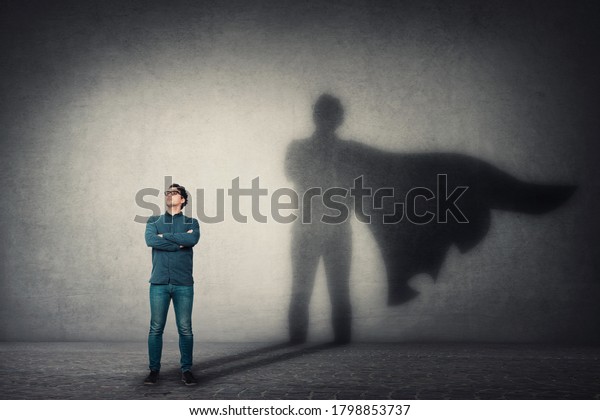 Brave man keeps arms crossed, looks confident,\
casting a superhero with cape shadow on the wall. Ambition and\
business success concept. Leadership hero power, motivation and\
inner strength symbol.