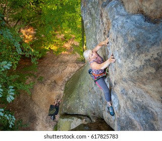 Brave Older Woman Rock Climber Is Climbing With Carbines And Rope On A Rocky Wall. Belayer Standing On The Ground Insuring The Climber. View From The Top. Extreme Sports Concept