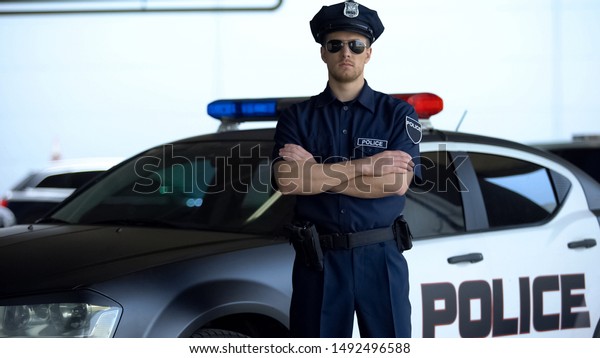 Brave police officer in service cap and\
sunglasses posing into camera near\
car