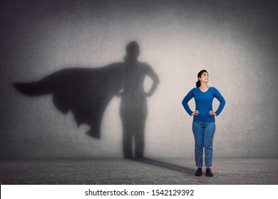 Brave Woman Keeps Arms On Hips, Smiling Confident, Casting A Superhero With Cape Shadow On The Wall. Ambition And Business Success Concept. Leadership Hero Power, Motivation And Inner Strength Symbol.