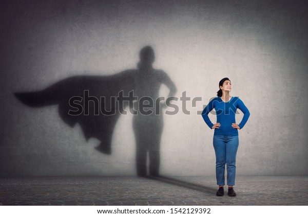 Brave woman keeps arms on hips, smiling\
confident, casting a superhero with cape shadow on the wall.\
Ambition and business success concept. Leadership hero power,\
motivation and inner strength\
symbol.