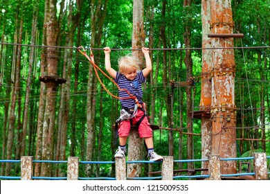 Brave Young Child In Safety Harness Climb High On Tree Tops, Pass Hanging Bridge Obstacle In Adventure Rope Park. Outdoor Activity,  Active Lifestyle In Summer Scout Camp On Family Holidays With Kids