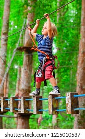 Brave Young Child In Safety Harness Climb High On Tree Tops, Pass Hanging Bridge Obstacle In Adventure Rope Park. Outdoor Activity,  Active Lifestyle In Summer Scout Camp On Family Holidays With Kids