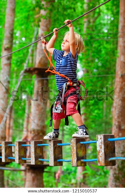 Brave young child in safety harness climb high on\
tree tops, pass hanging bridge obstacle in adventure rope park.\
Outdoor activity,  active lifestyle in summer scout camp on family\
holidays with kids