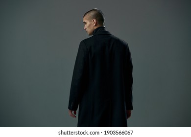 Brave young man dressed in black coat detective or criminal on gray studio background, back view