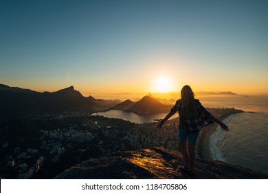 Brazil, Rio De Janeiro. Meeting The Dawn On The Hill Two Brothers. View Of The Big City, Lake, Ocean, Hills, Mountains. Orange-blue Colors Silhouette Of A Girl Who Looks At The City And Sun.