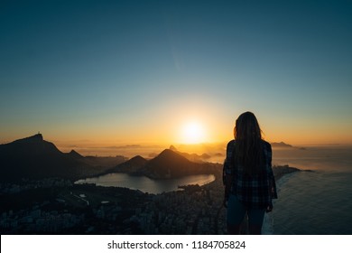 Brazil, Rio De Janeiro. Meeting The Dawn On The Hill Two Brothers. View Of The Big City, Lake, Ocean, Hills, Mountains. Orange-blue Colors Silhouette Of A Girl Who Looks At The City And Sun.