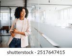 A Brazilian businesswoman smiles while looking to the side, standing in an office hallway that emphasizes a blend of professionalism and casual comfort. Casual professional lifestyle, business ease