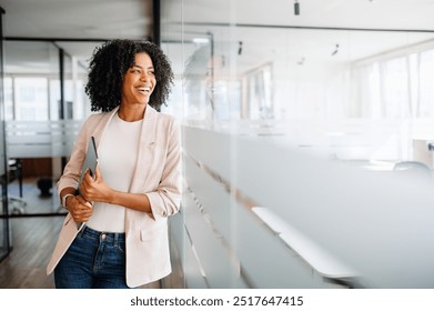 A Brazilian businesswoman smiles while looking to the side, standing in an office hallway that emphasizes a blend of professionalism and casual comfort. Casual professional lifestyle, business ease - Powered by Shutterstock