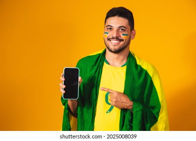 Brazilian Fan. Portrait, Brazilian Fan Showing His Cell Phone, Dressed As A Football Fan Or Soccer Match On Yellow Background. Colors Of Brazil. World Cup. 