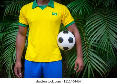 Brazilian Football Player In Brazil Flag And Colors Uniform Standing In Palm Frond Tropical Jungle Holding Soccer Ball