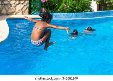 Brazilian Girl Jumping In Tropical Blue Swimming Pool In Summer.  Active Female Child Wearing Colorful Swimwear Outside In Pool Area