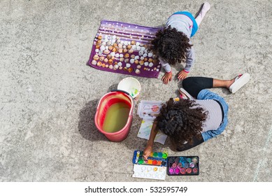 Brazilian Girls Painting Shells Outside On A Summer Day. Top View At Two Kids Sitting On A Patio Floor Painting With Watercolors, Image For Creative Family Concept, Hobby Blog, Toys  Business Website