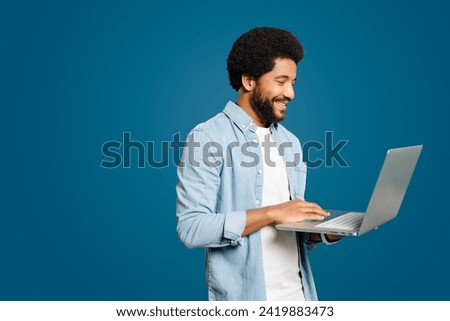 Brazilian man typing on the laptop isolated on blue. With a subtle smile and absorbed in his laptop, this man symbolizes the ease of accessing information and staying connected in modern digital world
