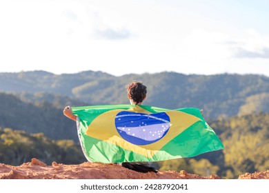 Brazilian seated atop a mountain, draped in the Brazilian flag, surrounded by a mountainous landscape. Brazilian supporter expressing support for Brazilian athletes in major sporting events - Powered by Shutterstock