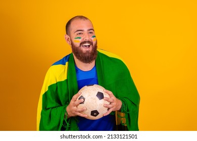 Brazilian Supporter Celebrating On Yellow Background Holding A Soccer Ball. Young Bearded Boy Big Fan Of Brazil