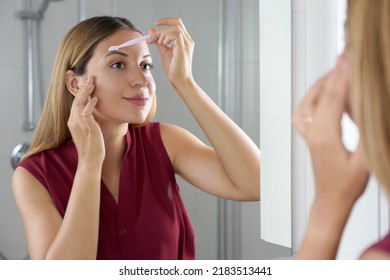 Brazilian woman shaving her eyebrows by razor at home. Pretty woman using razor on bathroom.
