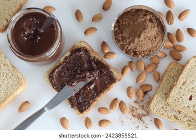 Bread slice with almond chocolate spread, a heavenly blend of rich almonds and smooth chocolate. Shot with many almonds, a bottle of almond chocolate spread and a bowl of coco powder in the background - Powered by Shutterstock