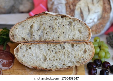 Bread Texture Of Home Baked Wheat Sourdough Bread Loaf With Seeds Cut In Half  With Smoked Ham, Salami, Grapes And Cherries On Wooden Board; Another Bread Loaf And Herbs In Background