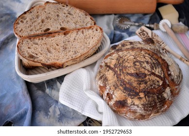 Bread texture of whole grain bread with flax seeds cut in half; dark bread sliced in wooden basket with another sourdough loaf of wheat bread on dish cloth and baking tools in background