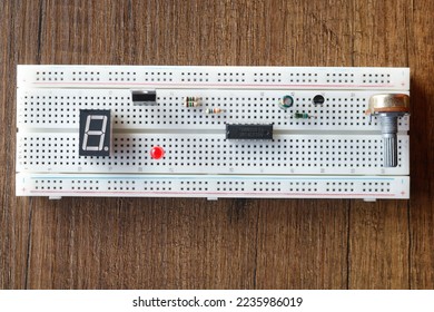 a breadboard with circuit elements on it. There are resistors, transistor, mosfet, Led, seven segment display, capacitor integrated and potentiometer on the breadboard with many holes. on wooden floor