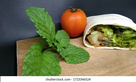 Breaded Chicken In A Tortilla Wrap With Lettuce And Tomato, With Mint Leave And Tomato As Properties. Isolated On Black Background