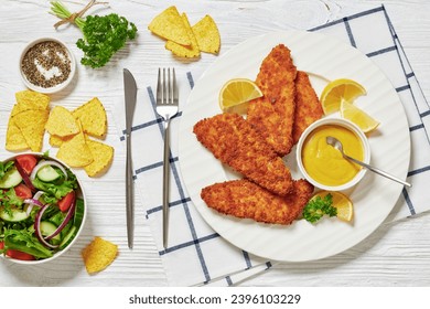 breaded fish fillet baked in oven served with yellow mustard on white plate, tortilla chips and fresh salad of lettuce, tomatoes, cucumber, red onions in bowl on white wooden table, flat lay - Powered by Shutterstock